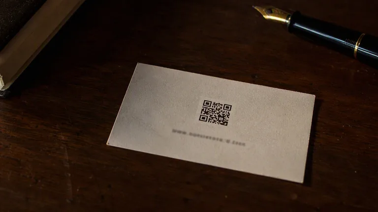 A business card lying on a dark wooden desk with a printed QR code on it, a fountain pen and notebook beside it