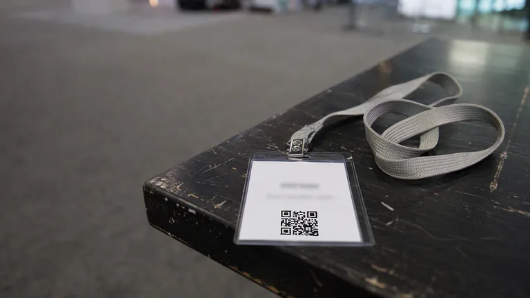 A conference lanyard badge lying face up on a table, its printed QR code showing, with the exhibition hall out of focus behind