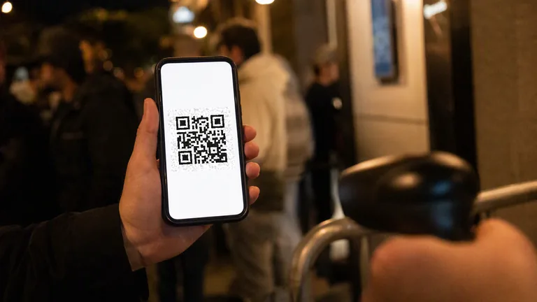 A hand holding a phone showing a QR code ticket at a venue entrance at night, with the queue out of focus behind