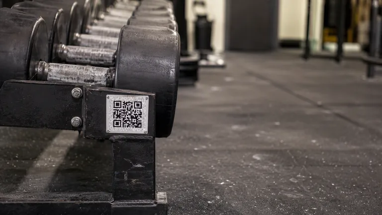 A small printed QR code plate bolted to the end of a dumbbell rack in a gym, with chalk dust across the rubber floor