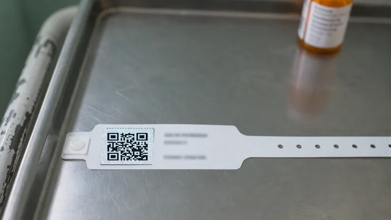 A printed QR code label on a stainless steel clinical tray beside a medication vial and a patient wristband