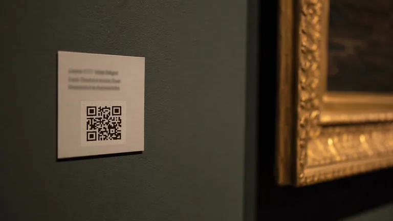 A printed gallery wall label carrying a QR code, mounted beside the gilt edge of a picture frame in dim museum lighting