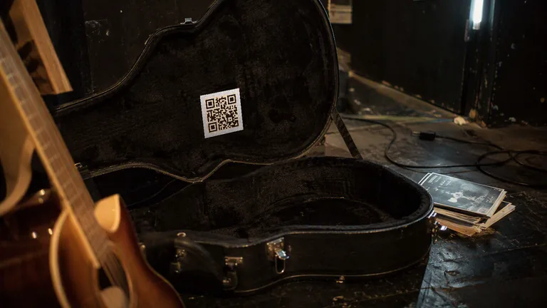 A printed QR code label inside the lid of an open guitar case backstage, with a stack of flyers on the floor beside it