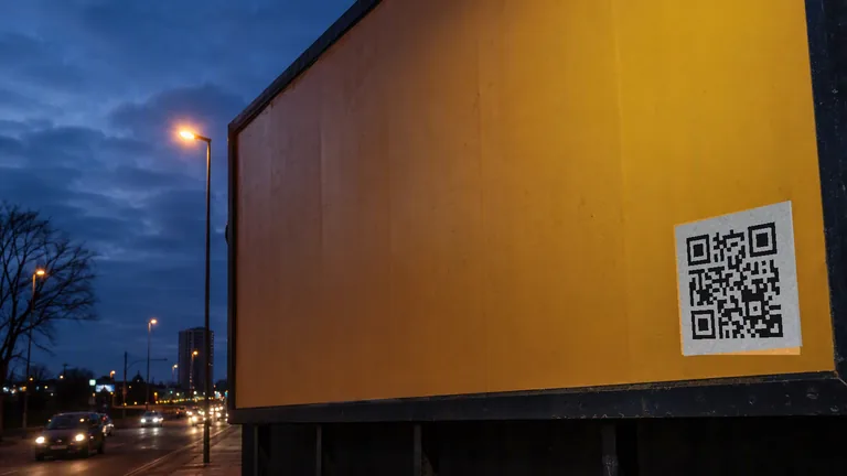 A large roadside billboard at dusk carrying a printed QR code panel, with traffic passing on the road below